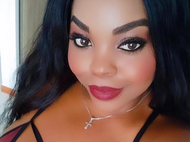 TatianaMarin has dark, wavy hair, brown eyes, and red lipstick, wearing a cross necklace with a black strappy top, and a slight smile showing her pouty ass.