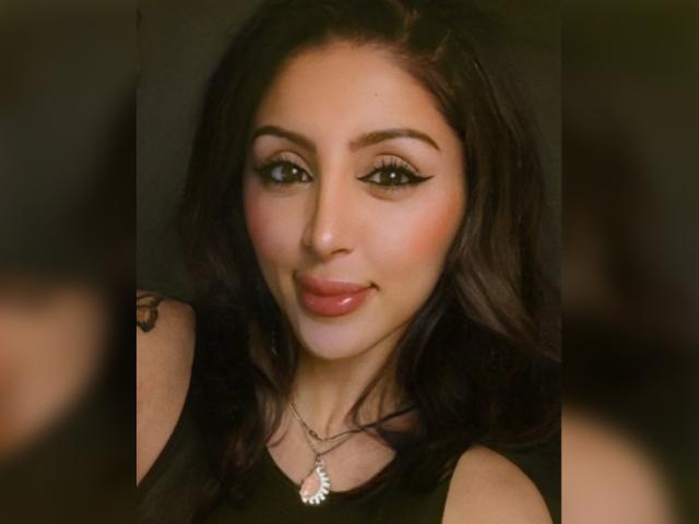 RaquelZ has dark wavy hair, green eyes with black eyeliner, pink glossy lips, and a black sleeveless top with a silver necklace featuring a small leaf design.