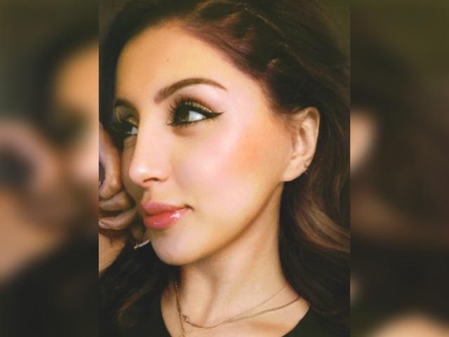 RaquelZ's face is a sexy, pretty, and cute ass with glossy pink lips, dark wavy hair, and bold eyeliner. She's wearing a black top and a gold necklace.