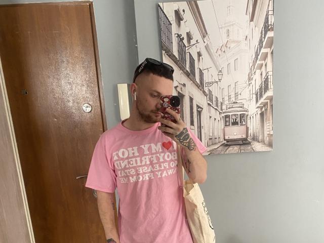 VityaKabura, with a pink t-shirt, tattoos on his right arm, and short buzzcut, takes a mirror selfie in a room, holding a beige bag and wearing white earbuds.