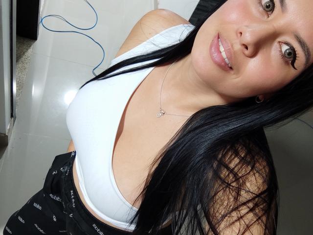 VenuzLove, con cabello negro largo, vistiendo una camiseta blanca, mostrando sus pechos, sonriendo con labios rosados brillantes, un collar de plata y pantalones negros en un selfie..