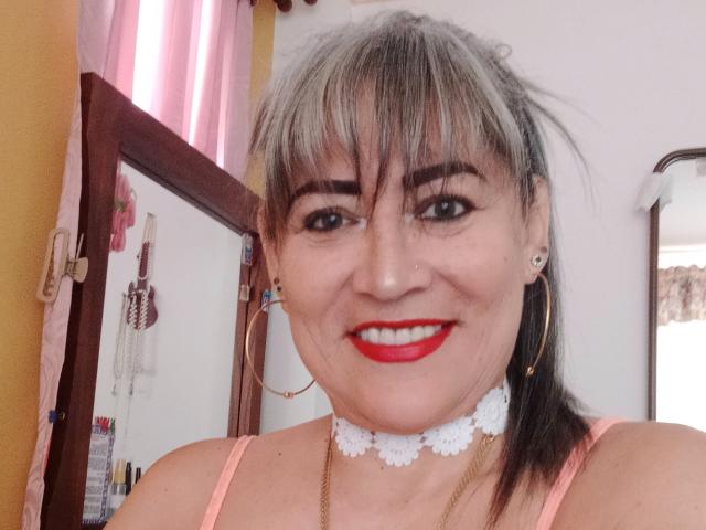 SamanthaLoveLatina with gray-streaked hair, red lipstick, white lace choker, gold hoop earrings, and pink top, smiling at the camera. Her tits are small, she's a Latina.