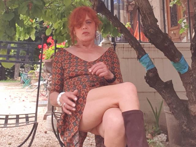 Lydye with red hair, wearing a patterned dress, sitting with one leg crossed, showing her bare thigh, and brown boots, under a tree with blue tape on the branch.