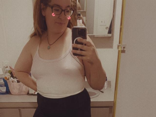 CountryCouple69 with glasses, white tank top showing her tits, black pants, taking a mirror selfie in a bathroom. Pink heart emojis on her face. Holding a phone.