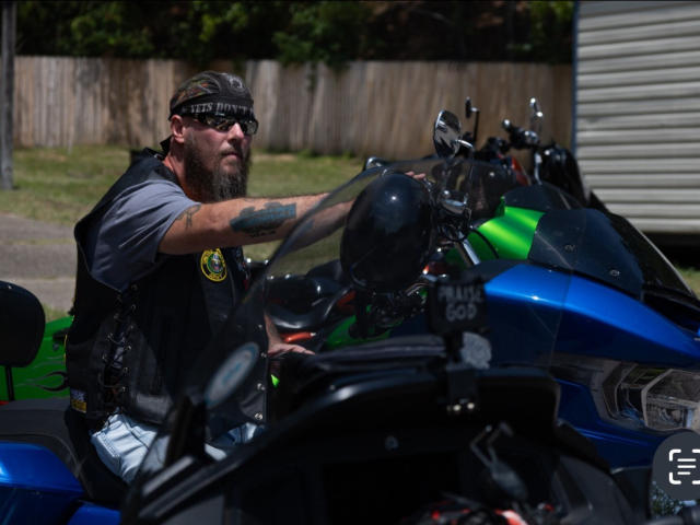 CountryCouple69, bearded biker with tattoos, wearing black vest and bandana, sits on blue motorcycle, gripping handlebars, green reflectors on bike, wooden fence in background, daytime, suburban setting.