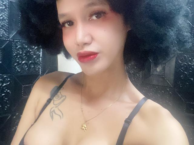 A photo of CumEaterJanna with short black curly hair, light brown skin, red lipstick, black bra, tattoo on left shoulder, gold necklace, and visible cleavage.