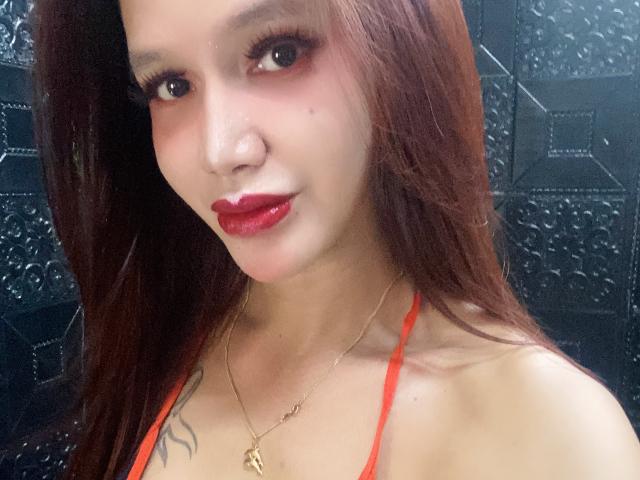 CumEaterJanna with long brown hair, red lipstick, and a red halter top, showing a small tattoo on her right shoulder and a gold necklace.