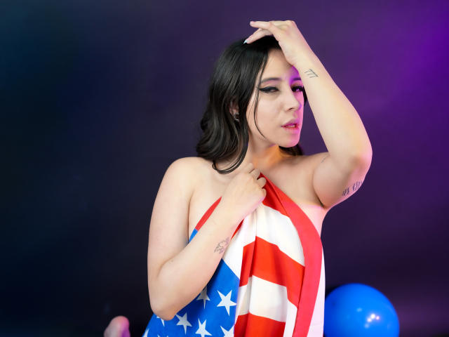 Kaiylin mit schwarzen Haaren, trägt ein einteiliges Kleid mit amerikanischer Flagge, bedeckt ihre Brüste mit ihrer Hand, im Hintergrund sind lila und blaue Ballons, sie hat ein Tattoo an ihrem rechten Arm..
