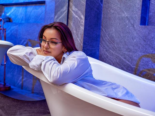 SalomeCastroXX, with glasses and purple hair, lounges in a white bathtub, wearing an oversized white shirt, exposing her bare ass, arms resting on the tub's edge.