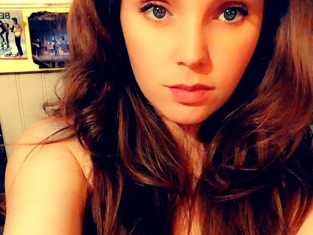 SquirtyNFlirty69 has long brown wavy hair, blue eyes, and is looking directly at the camera with a slightly pouty expression, wearing a sleeveless top.
