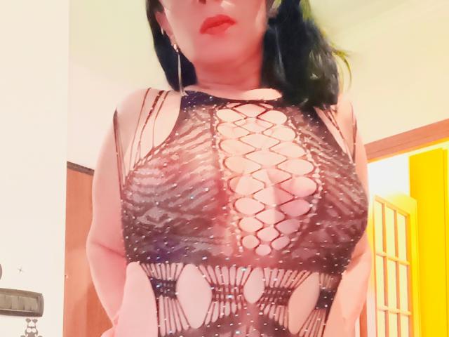 DivinaHot wearing a black, sheer, lace-up top that reveals her big boobs and nipples through the open pattern. Her red lips and black hair are partially visible.