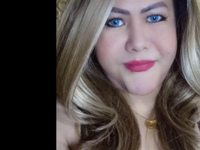 MistressBiancaS has blue eyes, pink lipstick, and blonde wavy hair with dark roots, wearing a gold necklace. Her ass is not visible.