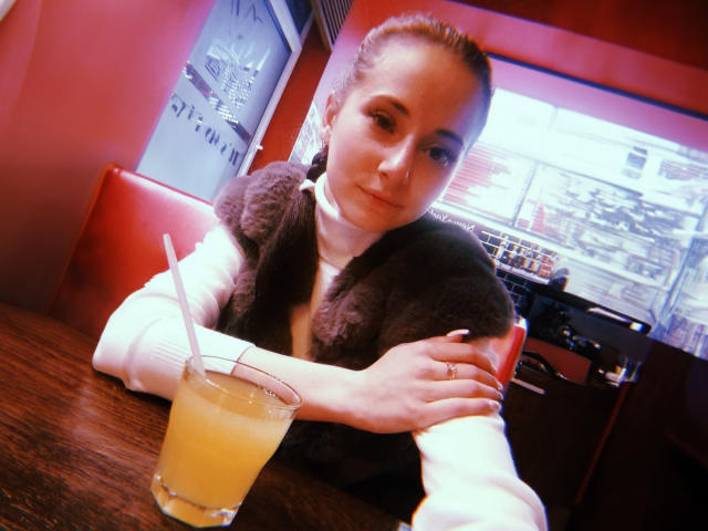 KrystalXMoon with brown hair in a bun, white turtleneck, black furry vest, arms crossed, drinking orange juice with a straw, sitting at a red diner table.