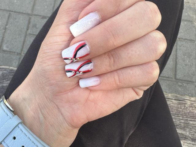 BellaJulls' hand with white, glittery nails, featuring red and black geometric designs on the index and middle fingers. Wearing a light blue wristband, black pants.