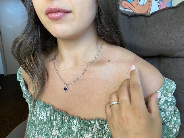 Mirellias Brüste sind leicht gerötet. Sie trägt ein grünes Oberteil mit Blumenmuster und einem Schulterauschnitt, eine silberne Halskette mit einem blauen Stein und einen Ring an ihrer rechten Hand, wobei ihre Lippen leicht geöffnet sind..