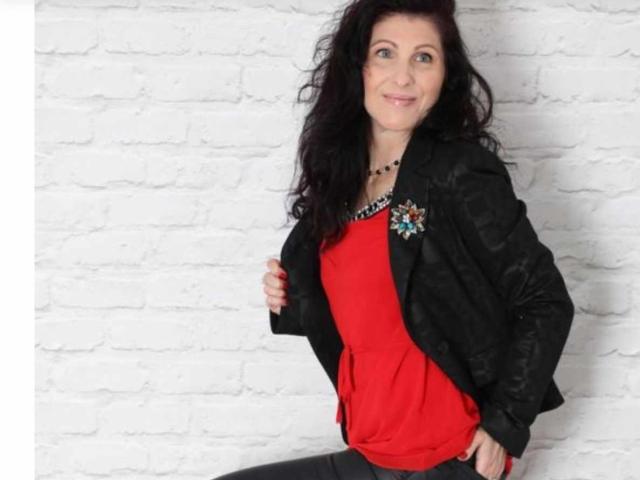 BettyLoveHot with dark wavy hair, red top, black jacket, and black necklace, smiling, hand on jacket, standing, white brick wall in background. She's looking cute and sexy.