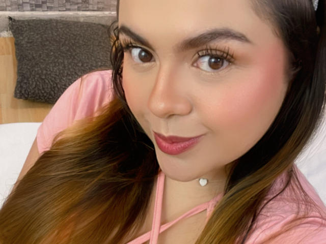 MissSofiia has pink lipstick, brown eyes, and long brown hair with ombre highlights. She's wearing a pink top and a heart-shaped necklace. Her ass is not visible.