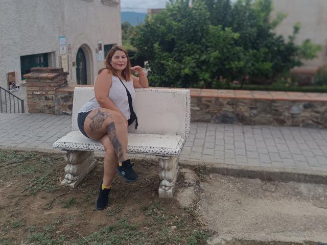 LouiseSexBigBoobs sitting on a white bench with a large tattoo on her right thigh, wearing a white top, black shorts, and black socks, holding a phone, smiling.