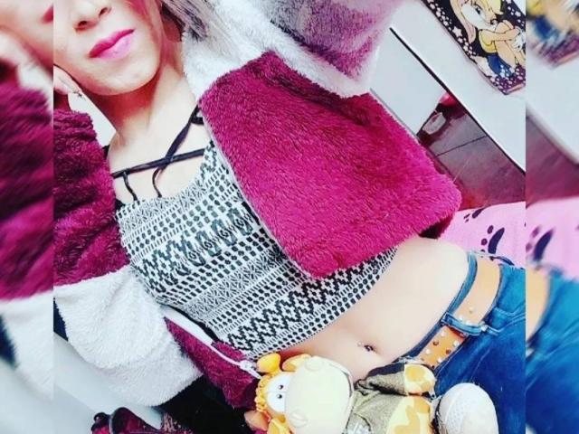 Lorenitha with pink lips, wearing a fuzzy red and white jacket, leopard print top, blue jeans, yellow belt, and stuffed toy, showing a navel piercing, in a selfie.