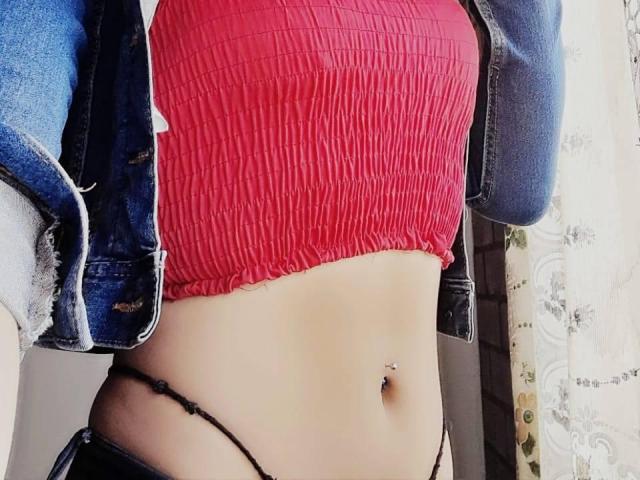 Lorenitha's red ribbed crop top, showing her tits, with a denim jacket, black thong, and belly button piercing, against a softly lit background.