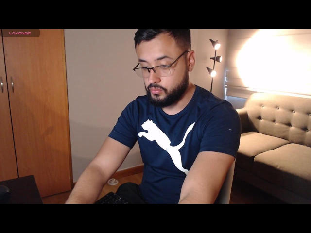 MassimoLee, short dark hair, beard, glasses, black Puma t-shirt, sitting, focused on laptop, room with wooden door, beige sofa, and lamp, looking slightly down, hands on keyboard.