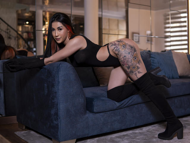 AnyaJay with black outfit, black gloves, and thigh-high boots, leaning on blue sofa, showing large tattoo on right thigh, and looking at camera with a slight smile.