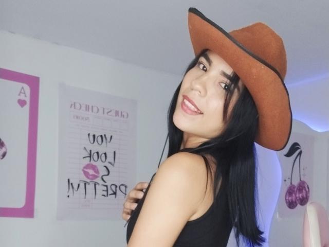 MichellAbi, avec de longs cheveux noirs, portant un chapeau de cowboy marron, un débardeur noir, souriant à la caméra, montrant ses fesses, avec du rouge à lèvres rose, des murs blancs et une jolie chambre..