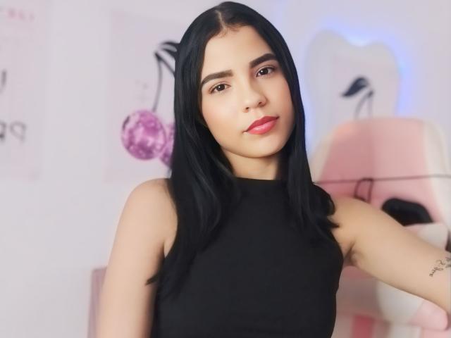 MichellAbi, avec ses cheveux noirs, porte un haut noir sans manches et on peut voir un tatouage sur son bras droit. Elle regarde l'appareil photo avec un léger sourire. Elle a du rouge à lèvres rose..
