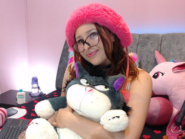 DandelionQueen avec un chapeau rose et duveteux, des lunettes et des cheveux bruns, tenant un peluche de chat gris et blanc, un oreiller en forme de licorne rose, sur un lit avec une couverture à motifs de cœur..