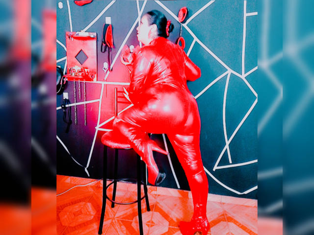 LexyDom in a shiny red latex suit, bent over a stool, gripping a wall with geometric patterns, wearing black heels, showing a round butt, with a red bag on the wall.