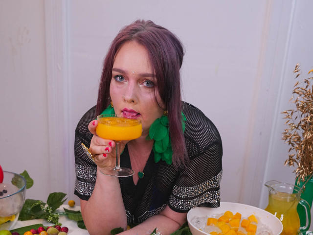 SinFulTitsDD with pink-tinted purple hair, green leaf earrings, and red lipstick, sips a colorful cocktail, wearing a black mesh top with white patterns, at a messy table.