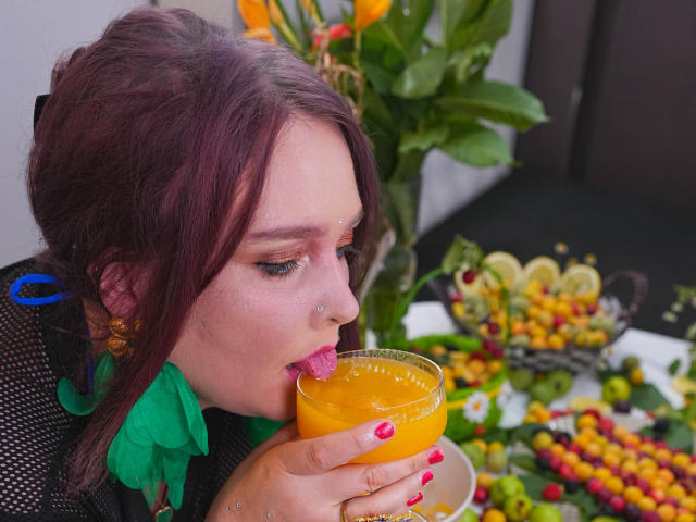 SinFulTitsDD with red hair, nose piercings, and green leafy collar, drinking orange juice, tongue out, holding glass, colorful fruit and plant in background, red nail polish.