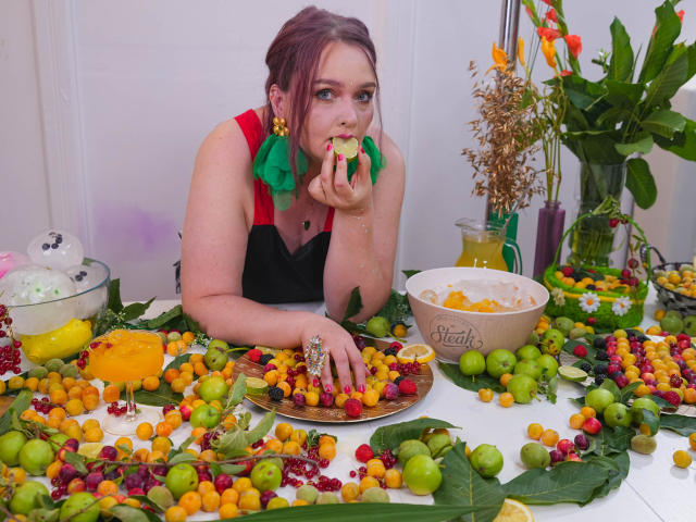 SinFulTitsDD with pink hair, eating grapes, has green and red earrings, black and red top, surrounded by colorful dried fruits and green leaves on a white table.