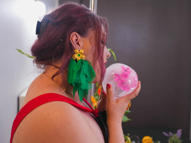 SinFulTitsDD with pink hair, wearing red and green floral earrings, blowing on a pink and white ice ball, showing off her red-straped back and red nail polish.