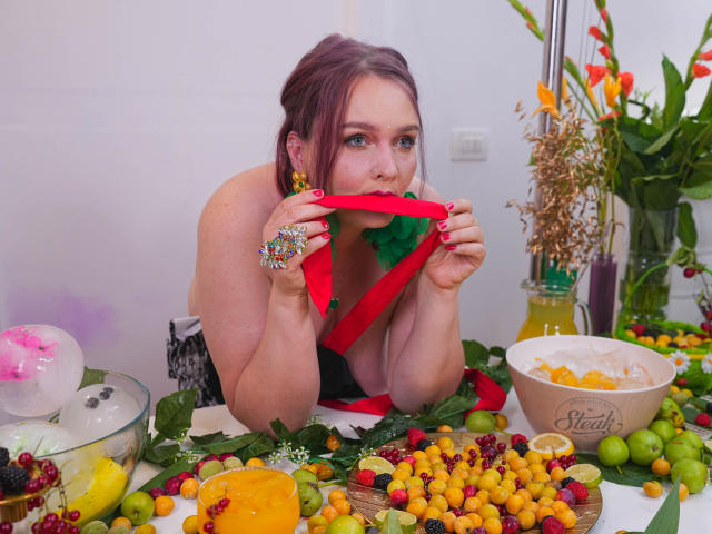 SinFulTitsDD with red panties in mouth, colorful ring on hand, green bra, red lips, holding green ribbon, surrounded by fruit, flowers, and bowl on white table.