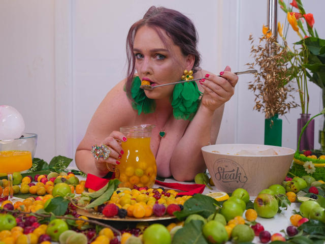SinFulTitsDD with green leaf earrings, eating grapes with a spoon, has orange juice in a glass, colorful fruit on a table, and a Teafit bowl in the foreground.