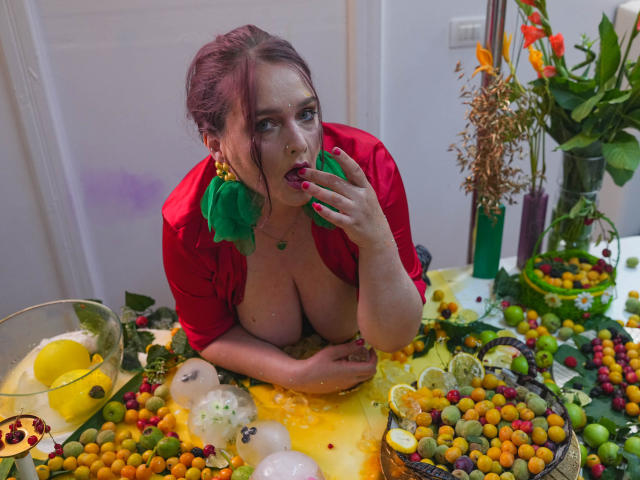 SinFulTitsDD with red dress, green earrings, and exposed tits, eating grapes, leaning over colorful fruit and lemon water, holding her tongue, in a bright, festive setting.
