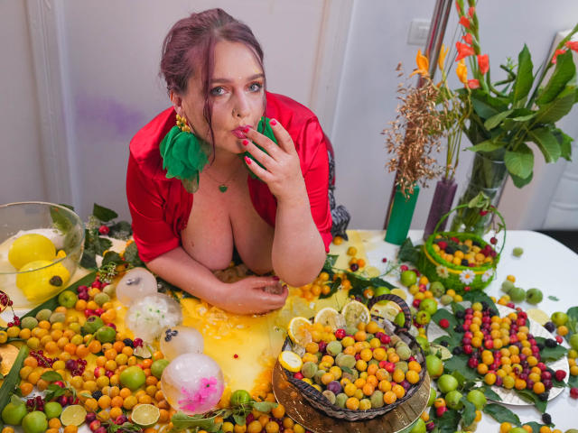 SinfulTitsDD with red shirt, green earrings, and lipstick, eating from yellow fruit tray, exposing big tits, colorful fruits, and lemon halves on white table, green leaf in mouth.