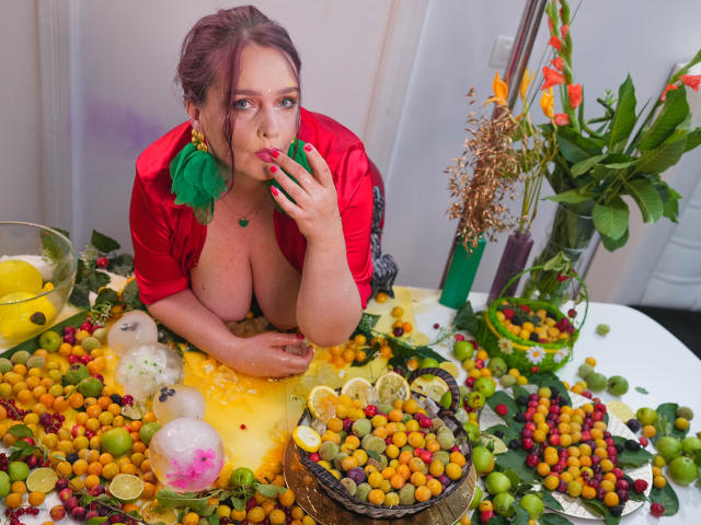 SinFulTitsDD with big boobs, red dress, and green earrings, eating a grape, surrounded by colorful grapes, lemons, and a basket on a white table with a vase.