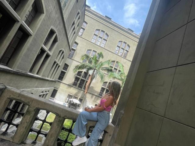 SashaPolet with tits in a pink top, ass in blue jeans, sitting on a concrete balcony, looking at a palm tree, urban building, and a blue sky.