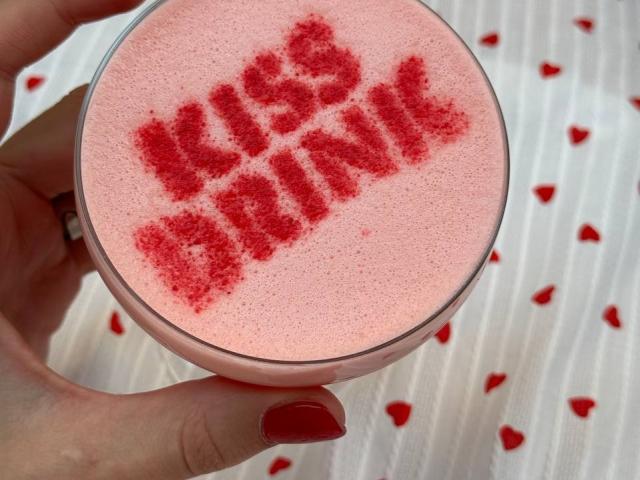 Tsukerberg's hand holds a pink, textured ice cream with red sugar sprinkles spelling "KISS ME" in the center. Her red-painted nails stand out.