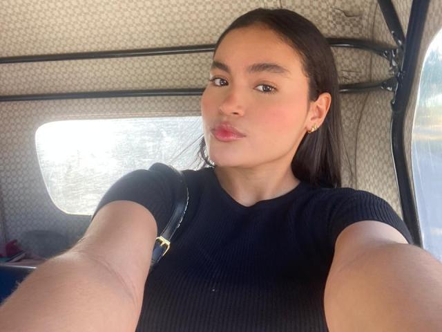 MiiaPalmer with dark hair, wearing a black ribbed shirt, taking a selfie in a car, showing a cute pout, small nose, and a gold earring, with a black strap.