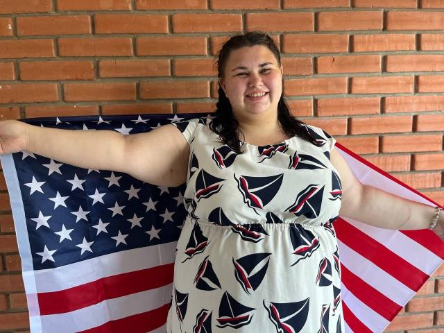 KaidoKun, chubby with dark hair in pigtails, smiling, wearing a white dress with triangle patterns, holds an American flag with arms out, brick wall background.