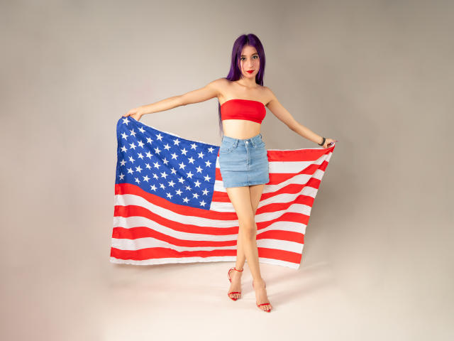 JuliaJobs com cabelo roxo, top de tubo vermelho, saia jeans azul, segurando uma bandeira americana, em pé, saltos vermelhos altos, mostrando o bumbum, magra, fofa, confiante..