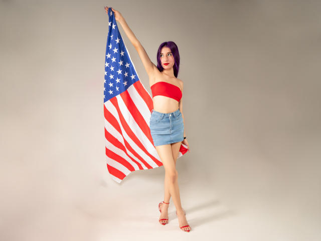 JuliaJobs with purple hair, red strapless top, and blue denim skirt, holding a large American flag, standing, red high heels, flag flowing, showing her ass.
