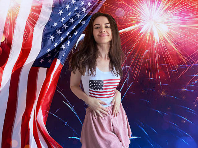 KaraCriegs with long brown hair, smiling, wearing a white tank top with a red/white American flag, pink pants, standing in front of a large American flag, fireworks in background.