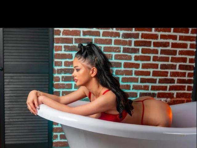 MolyStar with black ponytail, red bra, and red thong, leaning on white bathtub, showing her ass and small tits, against brick wall, looking to the side.