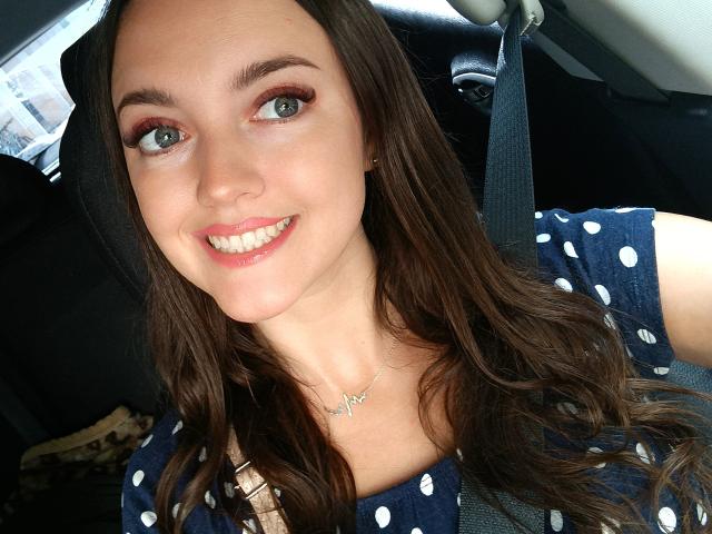 Vanilia69, with long brown hair and green eyes, smiling at the camera, wearing a blue polka dot dress, and a seatbelt, showing off her pearly white teeth.