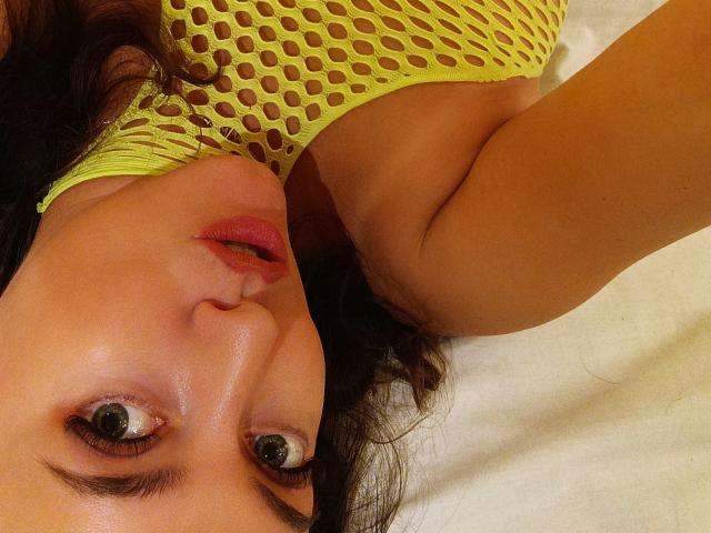 Vanilia69's face is up, wearing a neon yellow fishnet top, showing her big brown eyes, slightly parted pink lips, and brown wavy hair. Her tan skin is visible.