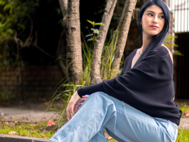EmaHenderson met donkerblauw haar, een zwarte sweater met een open schouder, en lichtblauwe broek, zittend buiten, naar de zijkant kijkend, met roze lippenstift, een slank postuur, kleine borsten, en een subtiele glimlach..